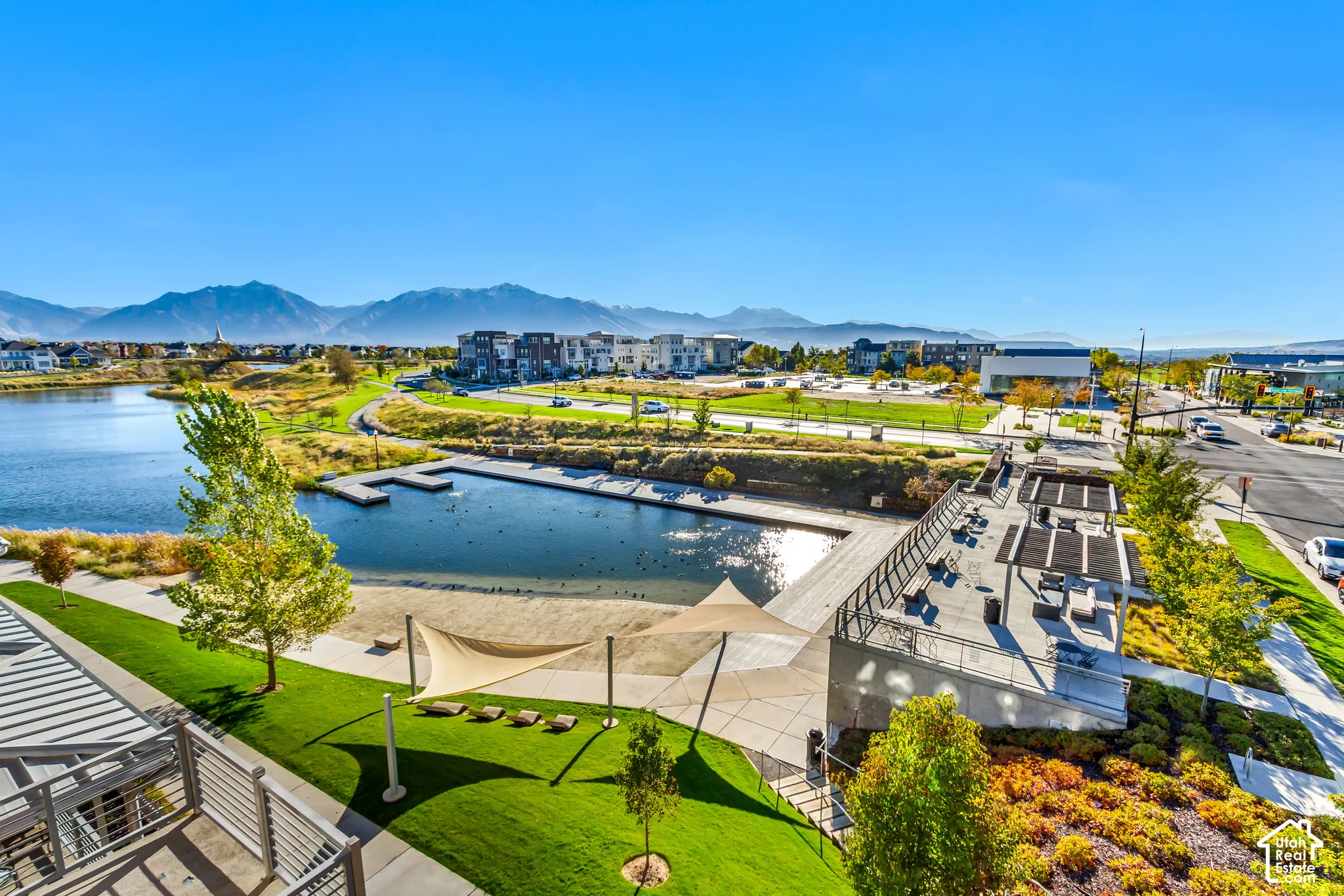 Elevated outdoor patio with overhead pergola, concrete hardscaping, manicured lawn, water feature with dock, and prominent mountain backdrop