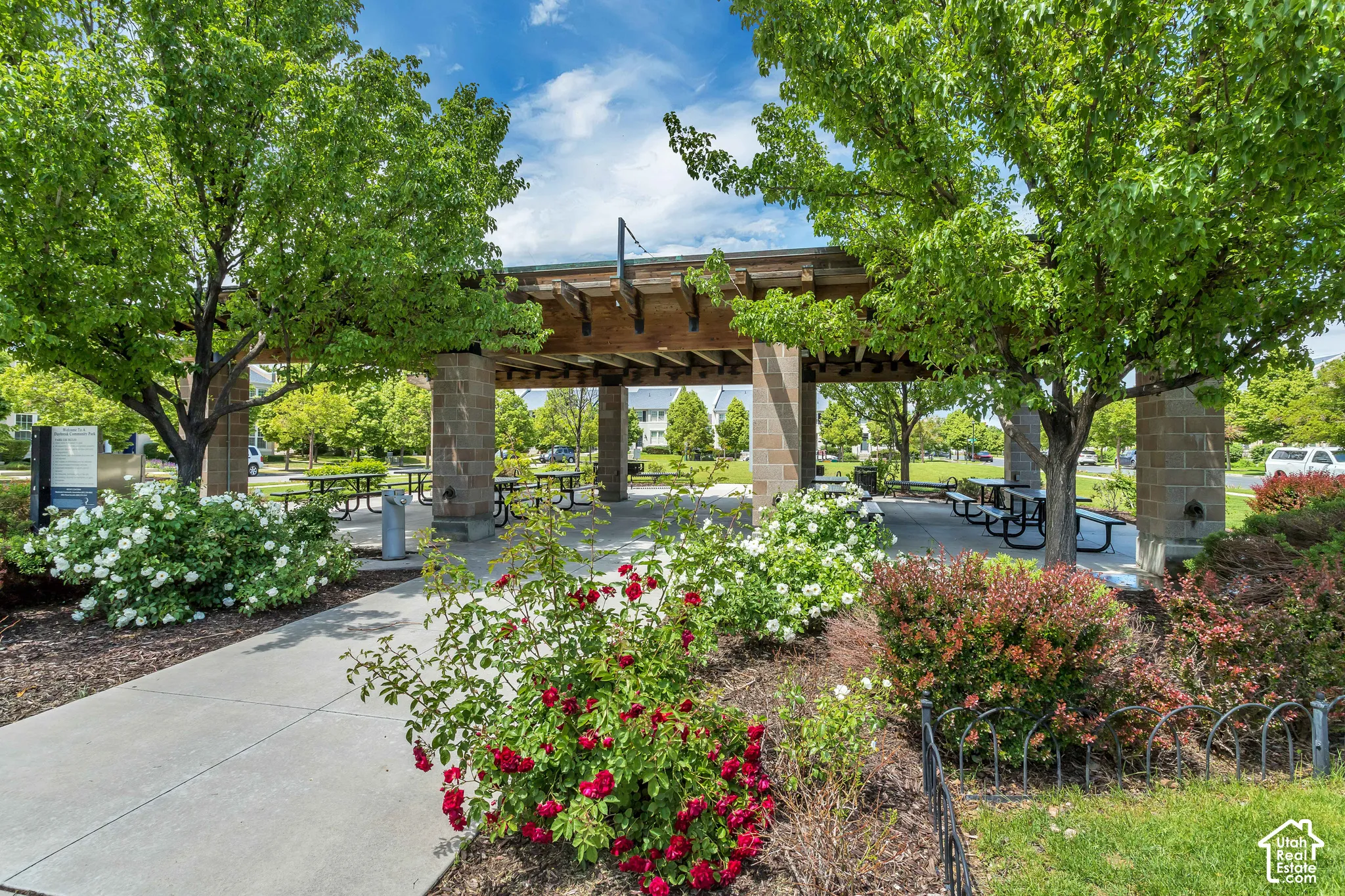 Open-air pavilion featuring a timber-beam pergola, stone masonry pillars, and a concrete patio surface, surrounded by mature deciduous trees and flowering landscape shrubbery