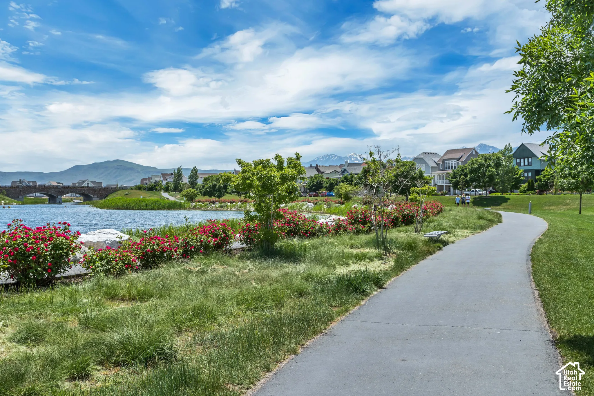 Paved walking path bordering a lake, featuring stone masonry bridge architecture, mountain backdrop, and landscaped floral vegetation