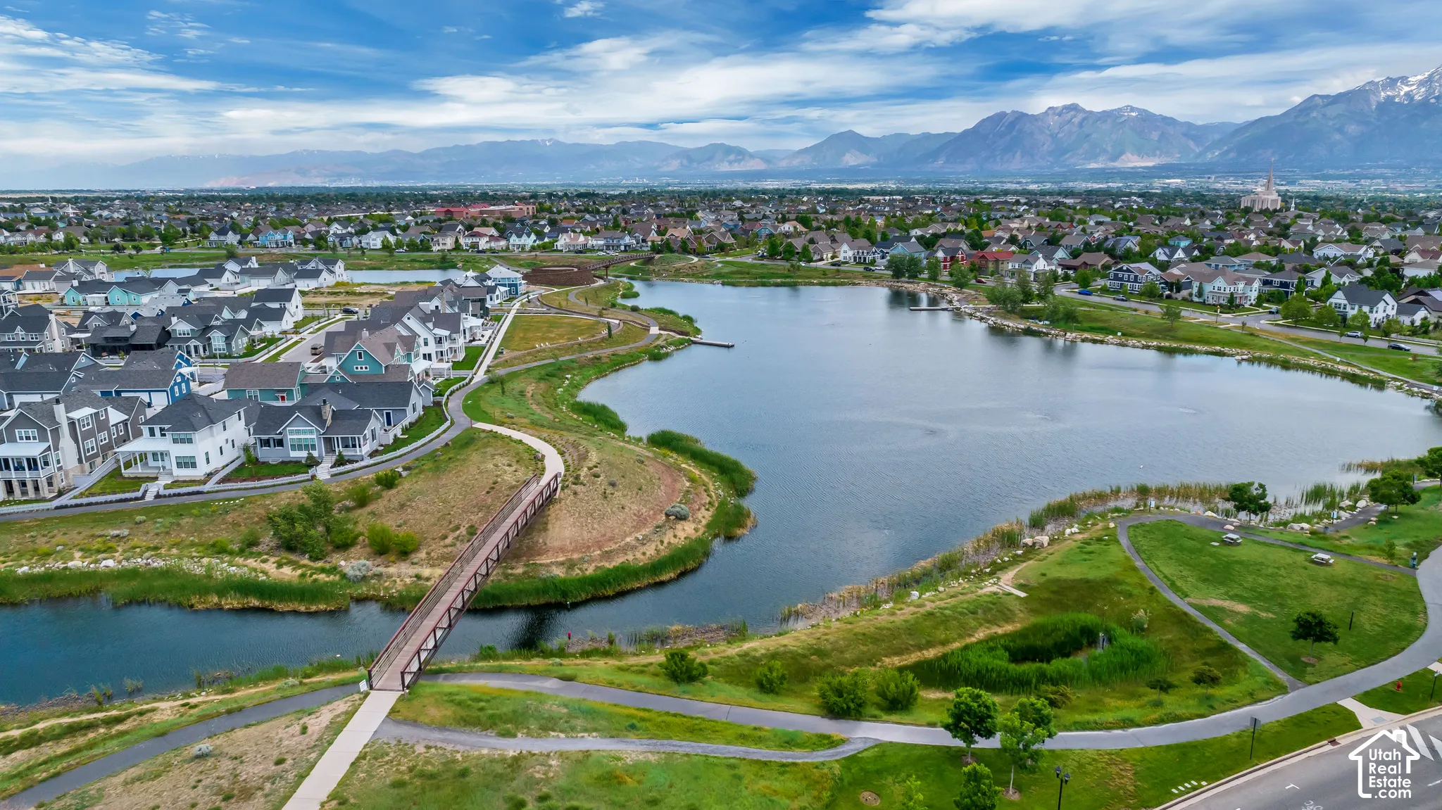 Aerial perspective featuring a large lake, pedestrian bridge, paved pathways, and mountain backdrop