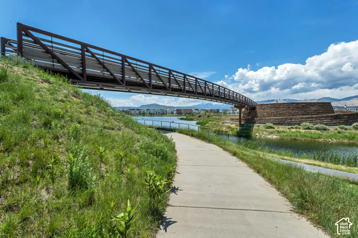 Pedestrian bridge featuring metal railings, concrete path, pond, and stone-retaining wall structures set against a mountain backdrop
