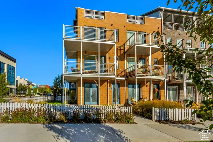 Multi-level brick exterior featuring stacked balconies with metal railings, glass-paneled double doors, and a perimeter picket fence