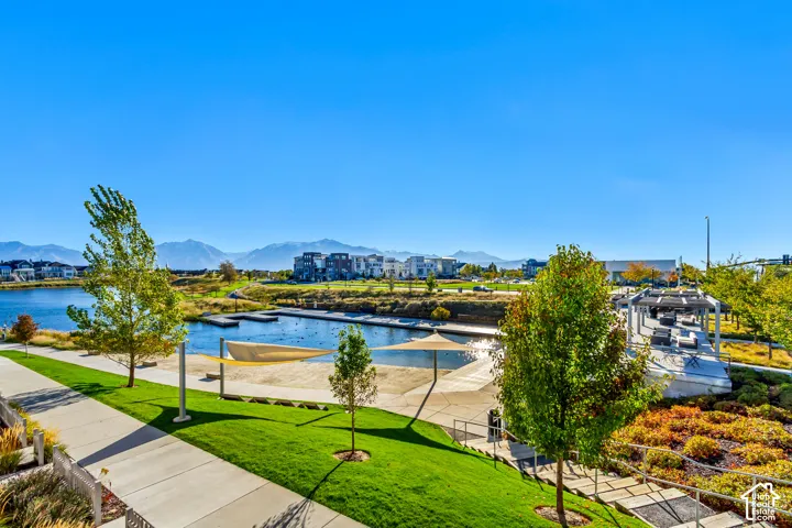 Waterfront community area featuring a pond, paved pedestrian paths, landscaped grounds with mature trees, and a mountain backdrop