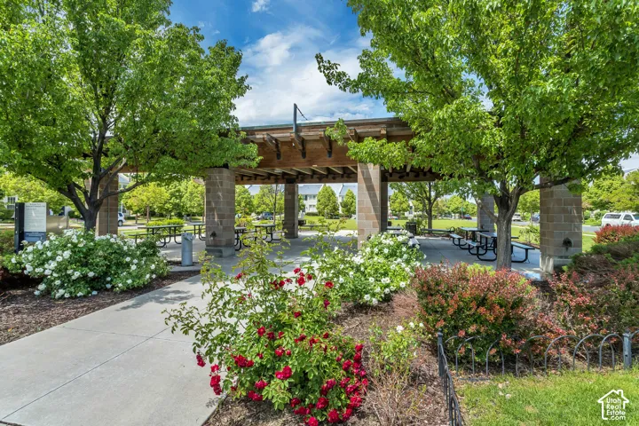 Open-air pavilion featuring a timber-beam pergola, stone masonry pillars, and a concrete patio surface, surrounded by mature deciduous trees and flowering landscape shrubbery