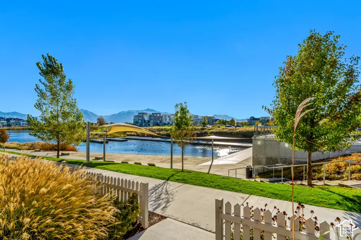 Waterfront parkland featuring a sandy shoreline, concrete pedestrian pathways, shade structures, and manicured lawns against a mountain backdrop