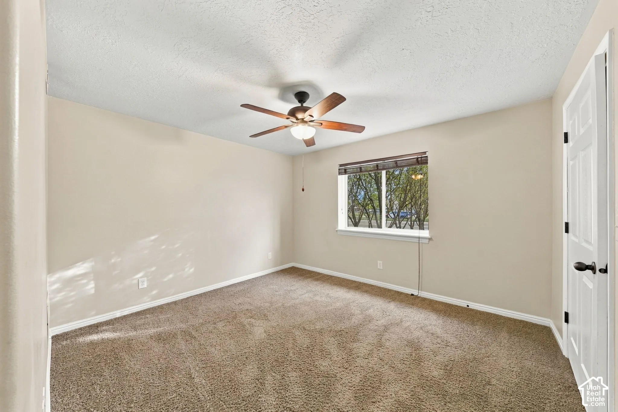 Neutral-toned interior space featuring wall-to-wall carpeting, a ceiling fan with integrated lighting, and a window with wood-finish blinds