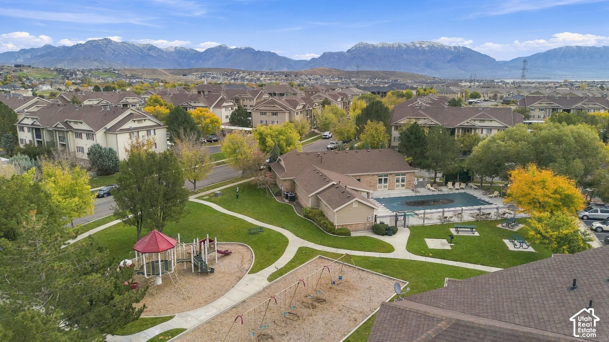 Community recreation area featuring a swimming pool, fenced playground, mature trees, and mountain scenery