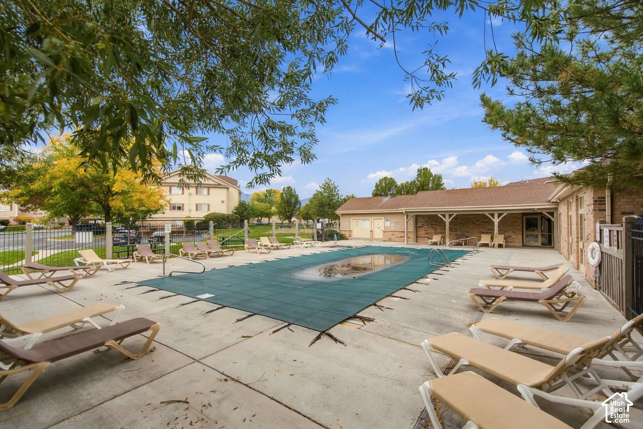 Swimming pool with safety cover, expansive concrete patio, covered outdoor area with support columns, and perimeter metal fencing