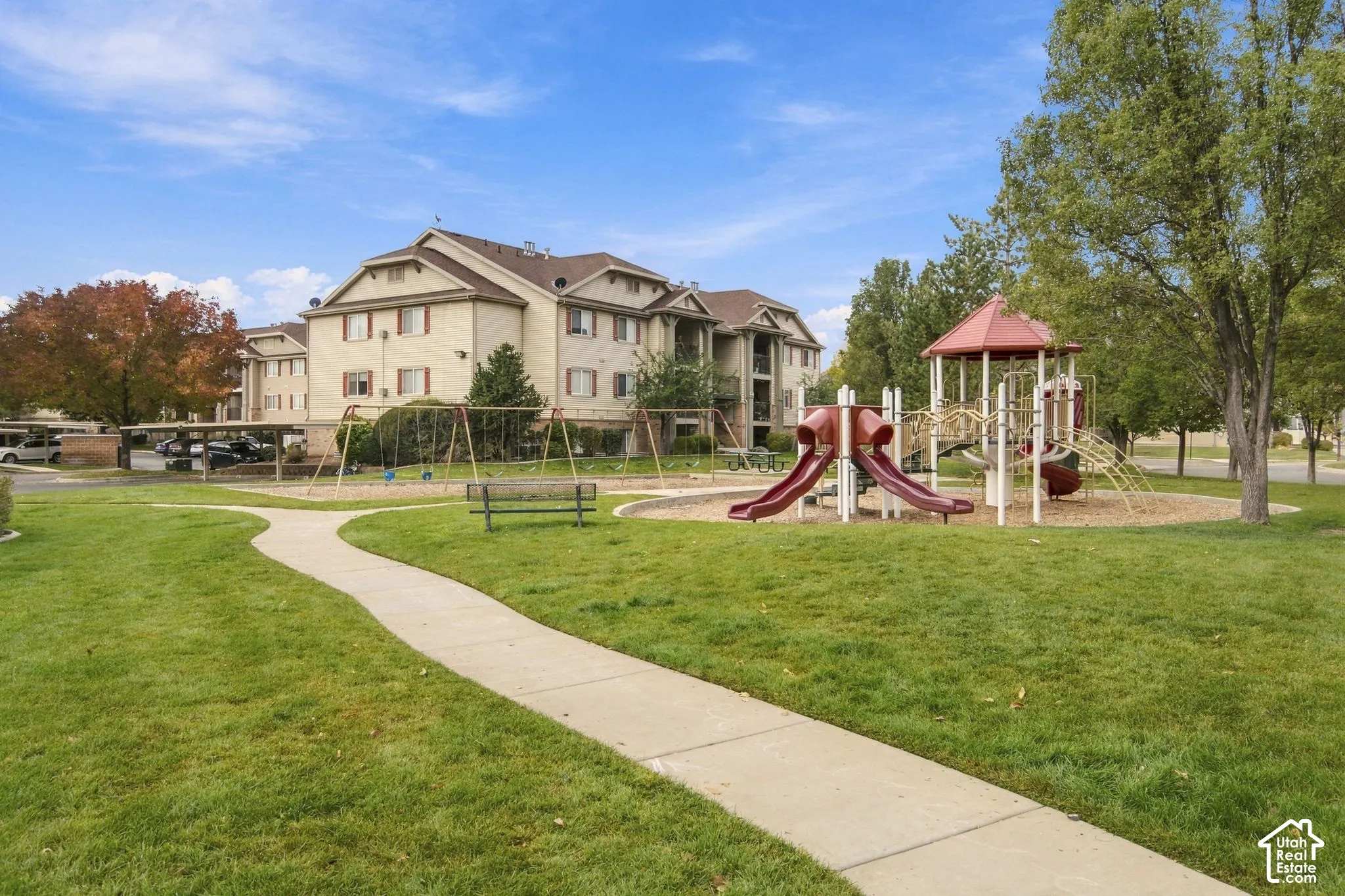 Common area featuring a paved walkway, playground equipment, and an exterior bench situated on landscaped grounds