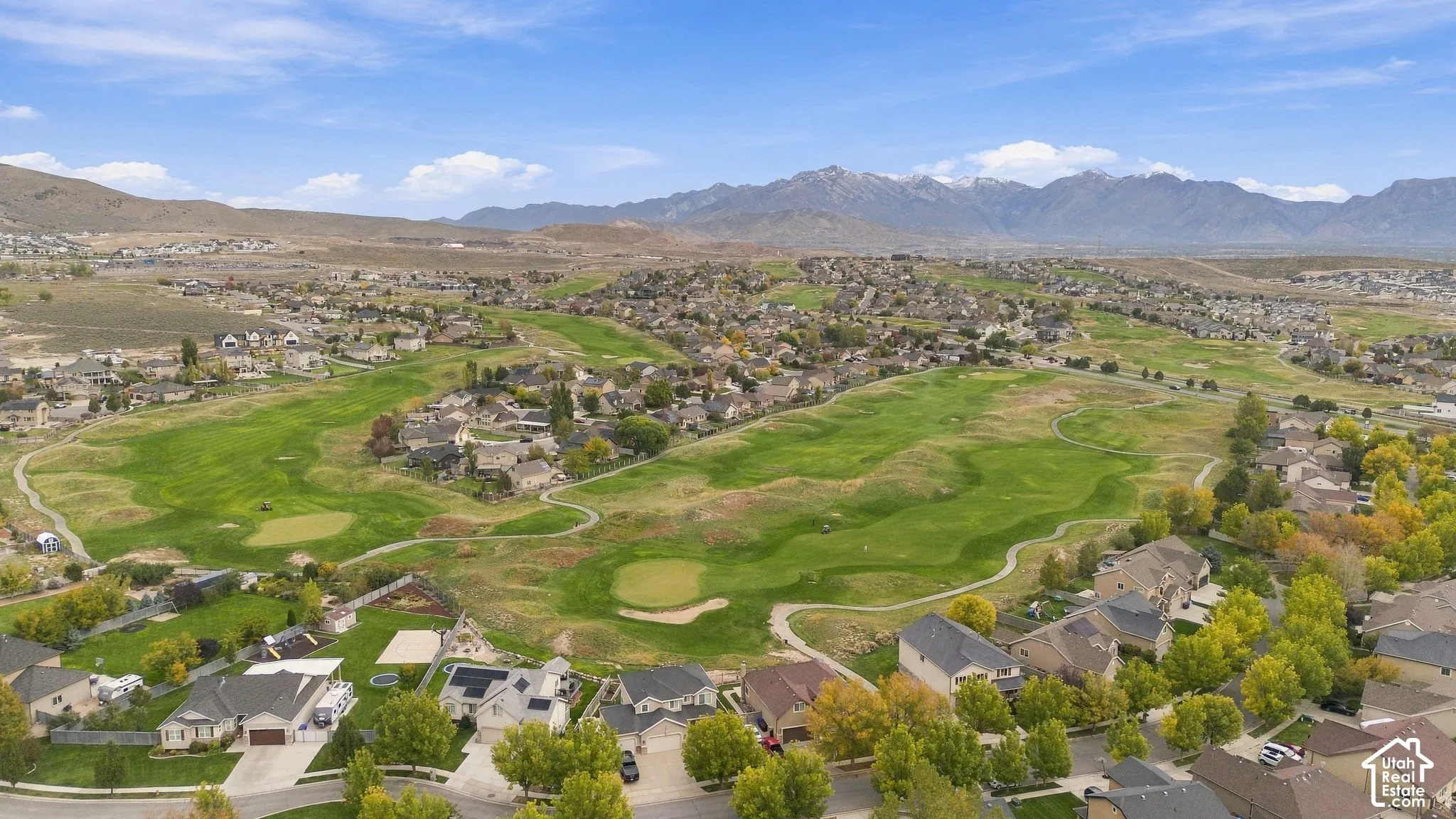 Aerial perspective of a residential community featuring extensive green space, manicured golf course fairways, paved paths, and mountain backdrops