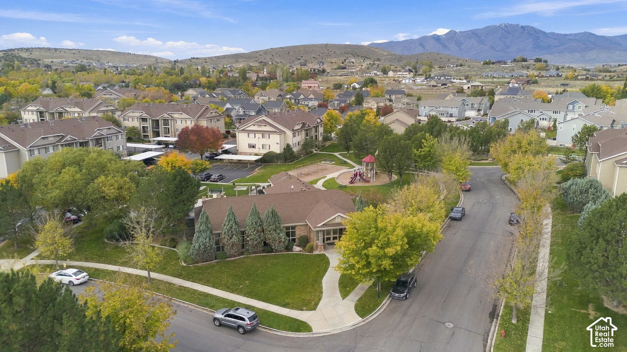 Aerial perspective featuring an asphalt-shingled residential structure, mature landscaping, paved walkways, a playground area, and a backdrop of mountainous terrain