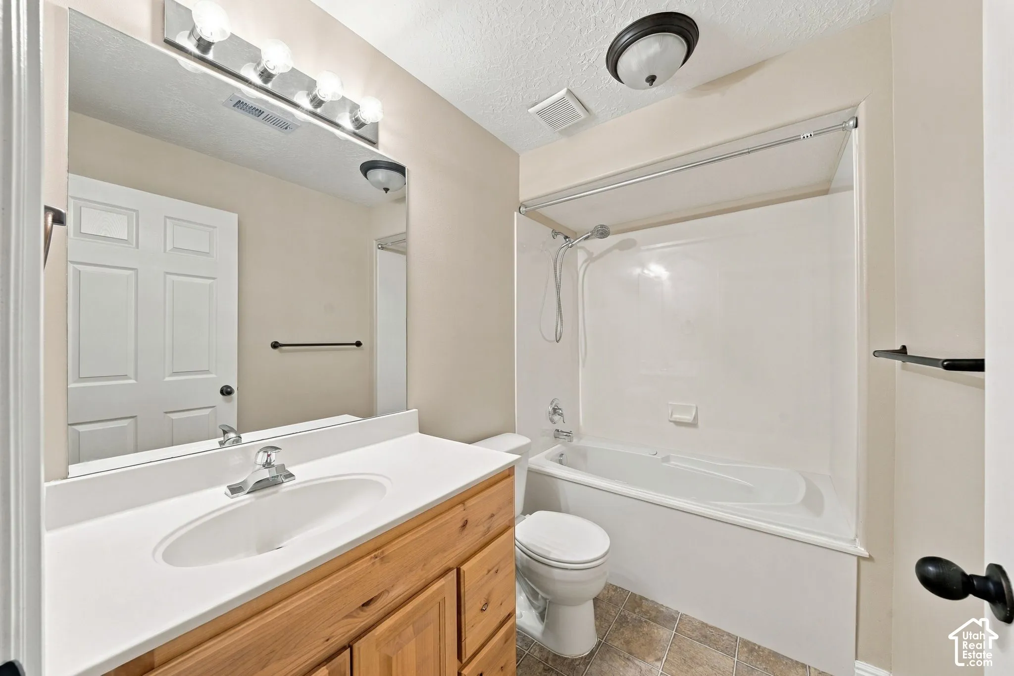 Bathroom featuring a wood-finish vanity with a white countertop, oval drop-in sink, wall-spanning mirror, and a shower-tub combination with a handheld showerhead fixture