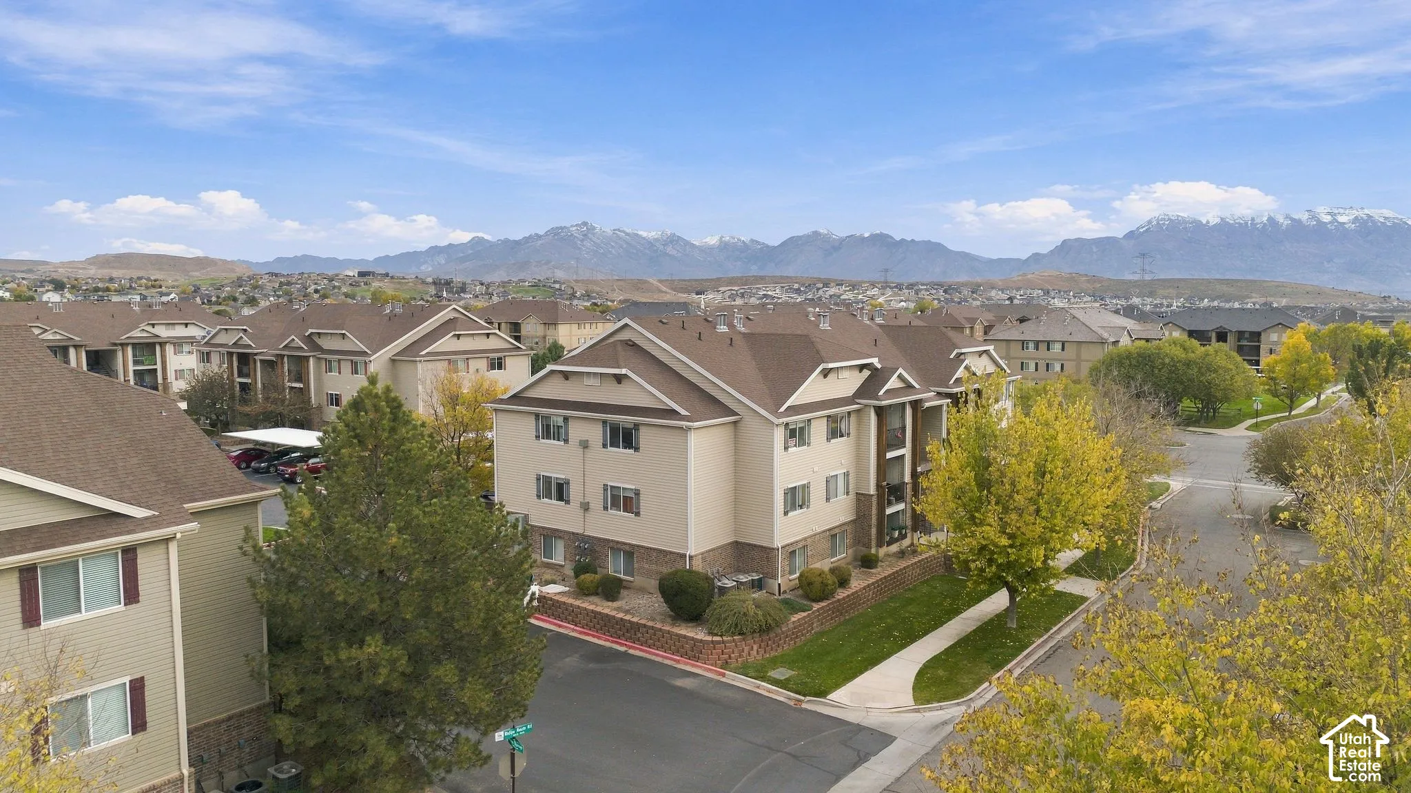 Multi-story residential buildings featuring tan siding, brick masonry detailing, asphalt shingle gable roofs, and mountain range backdrop