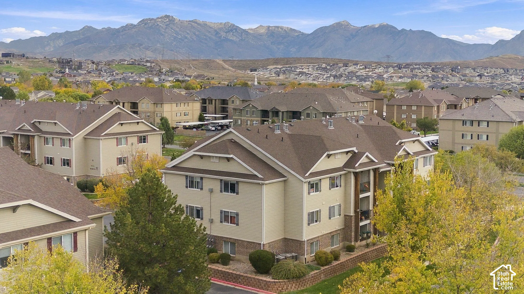 Multi-story residential buildings featuring tan horizontal siding, brick veneer foundations, asphalt shingle rooflines, and mountain backdrops