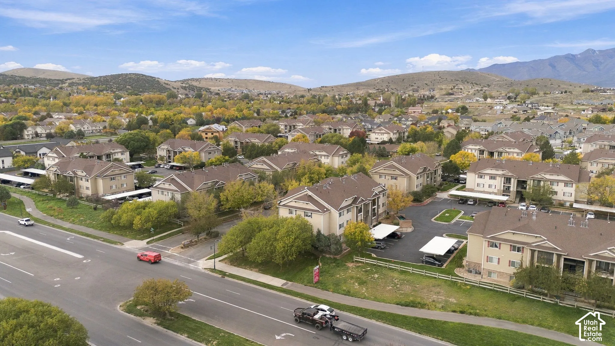 Multi-building residential complex featuring pitched roofs, neutral-tone stucco exteriors, paved surface parking lots with covered carports, and mature landscaping against a mountain backdrop