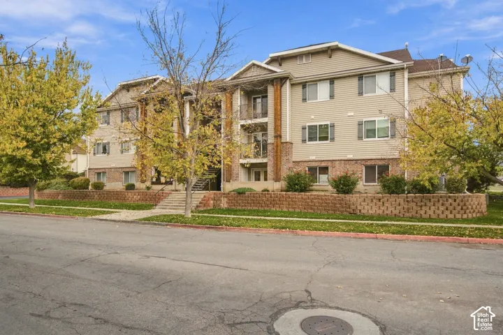 Multi-story exterior featuring tan horizontal siding, brick veneer accents, multi-level balconies with wood-finish supports, and a segmented stone-block retaining wall