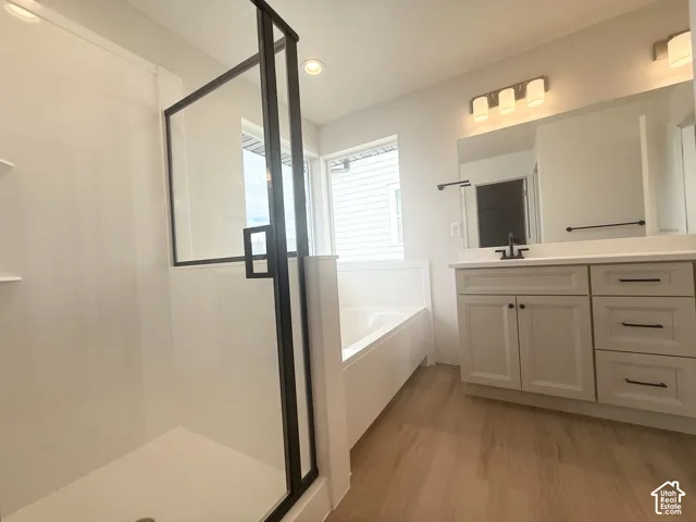 Bathroom featuring a walk-in shower with black-frame glass enclosure, a white built-in soaking tub, a vanity with neutral-toned cabinetry, light-colored stone-surface countertop, and wood-finish flooring