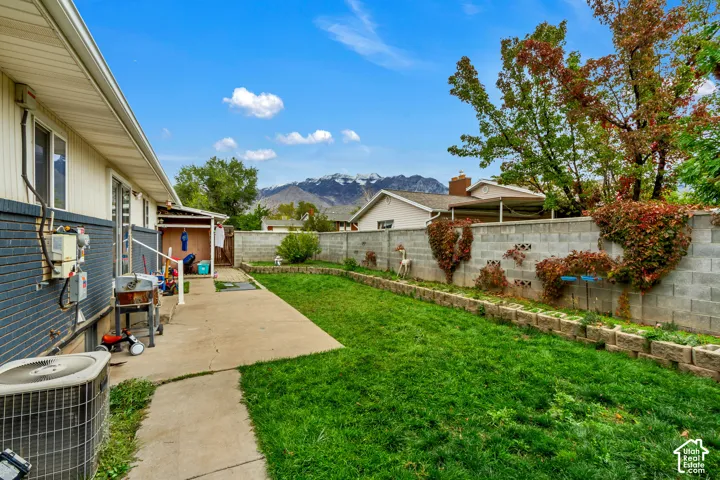 Fenced backyard featuring a concrete patio, tiered stone retaining walls, manicured lawn, and a mountain backdrop