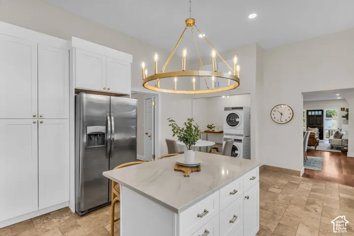 Kitchen featuring white shaker-style cabinetry, a central island with a solid surface countertop, stainless steel refrigerator, stone tile flooring, and a decorative circular chandelier