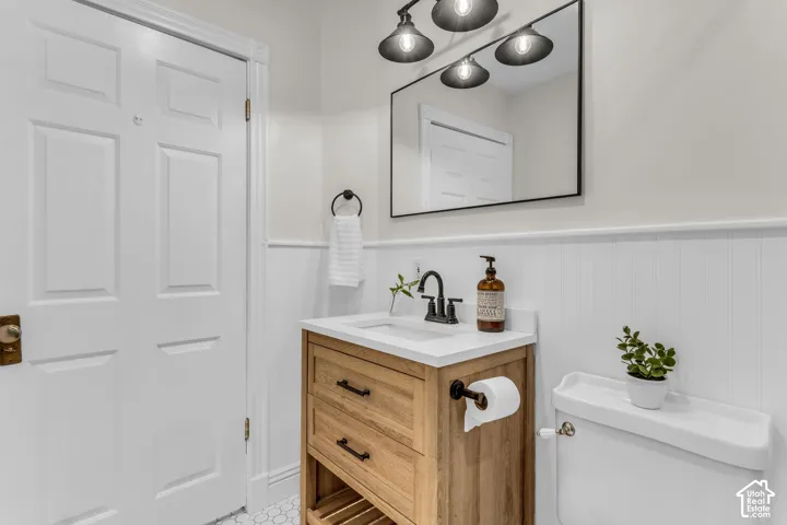 Bathroom featuring a wood-finish vanity with a stone countertop, white beadboard wainscoting, a rectangular vanity mirror with a black frame, matte black hardware, and three-light industrial-style wall lighting