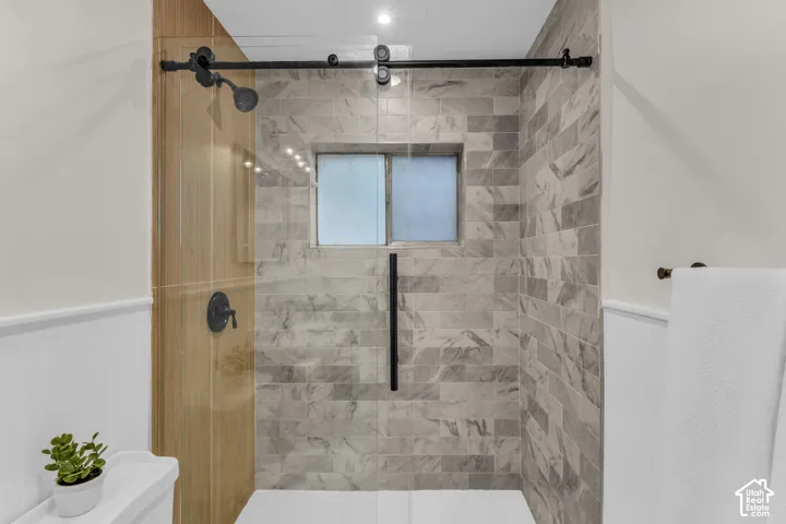 Walk-in shower featuring a glass enclosure with a black sliding track system, matte black rainfall shower head, light-toned stone-finish tile surround, and wainscoting