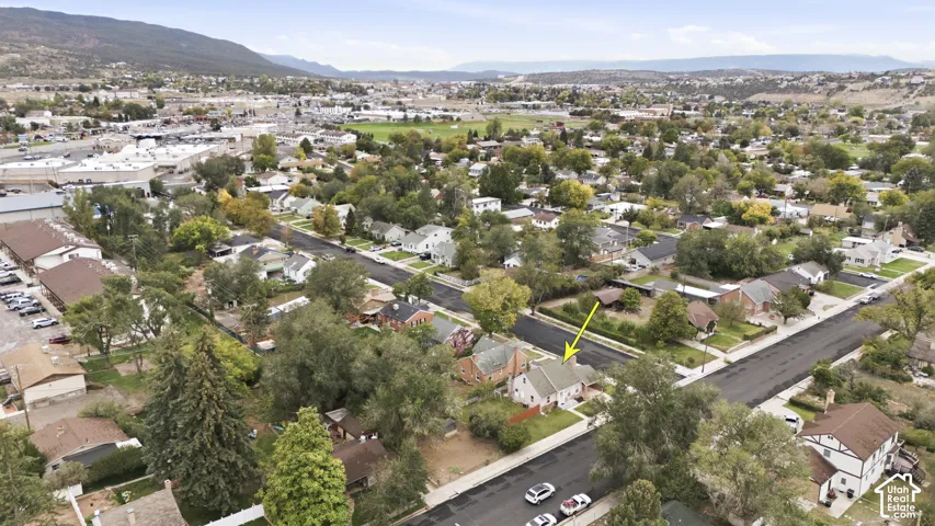 Aerial perspective of a detached residential property situated on a corner lot, featuring a pitched roofline, mature trees, and mountainous terrain in the distance