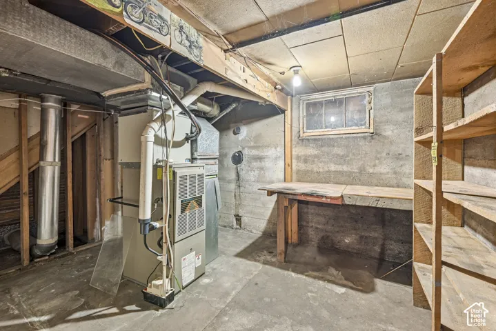 Unfinished basement space featuring exposed concrete walls, a high-efficiency furnace, a built-in wooden workbench, and dedicated wood-framed shelving units
