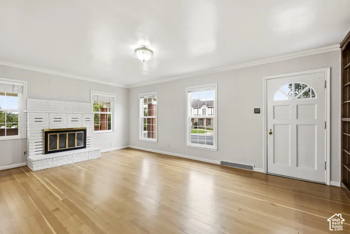 Living area featuring light wood-finish flooring, a white-painted brick fireplace, crown molding, multi-pane windows, and a classic-style entry door with a decorative transom window