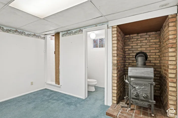 Cast-iron wood stove featuring a brick surround and hearth, drop ceiling, and carpeted flooring