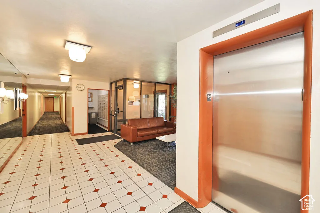 Building lobby featuring a brushed-metal elevator with a wood-toned frame, tiled flooring with geometric accents, full-length wall mirror, and integrated mailboxes