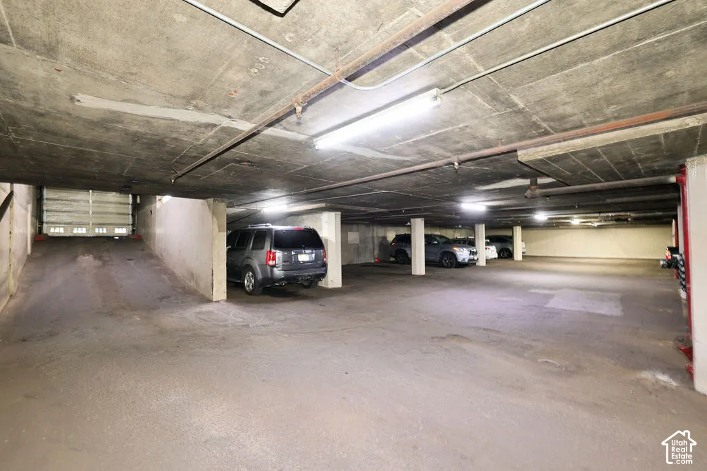 Enclosed parking garage featuring concrete flooring, structural pillars, overhead lighting fixtures, and a secure ramped entrance
