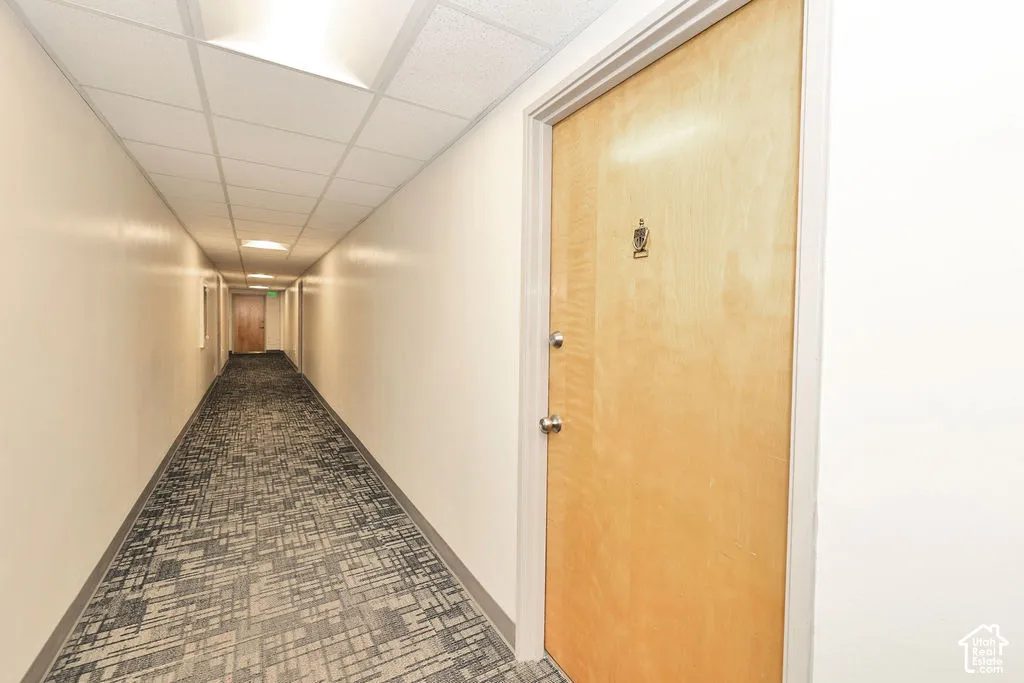 Hallway featuring a grid-patterned carpet, smooth-finish walls, a drop-ceiling system with recessed lighting, and a flush-panel wood-finish door with brushed metal hardware
