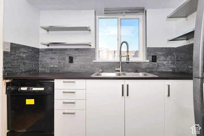 Kitchen area featuring white cabinetry, dark countertop, dual-basin stainless steel sink with high-arc pull-down faucet, stacked stone backsplash, and stainless steel floating shelving