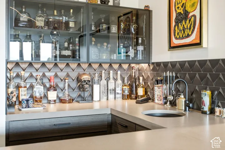 Wet bar featuring flat-panel cabinetry, glass-front upper storage, solid surface countertops, a geometric tile backsplash, and a matte black faucet