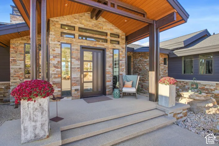 Contemporary exterior featuring natural stacked-stone masonry, prominent wooden ceiling beams, a recessed front entry with vertical window panels, and integrated concrete steps