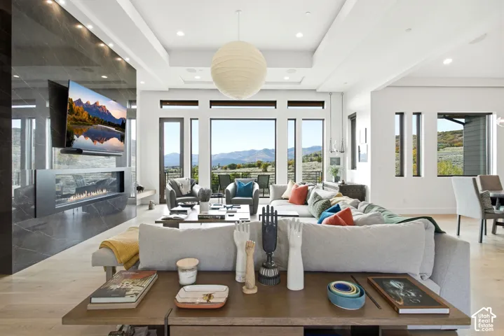 Open-concept living space featuring high tray ceilings, large-format tile fireplace surround, linear gas hearth, wood-finish flooring, and expansive windows with mountain scenery