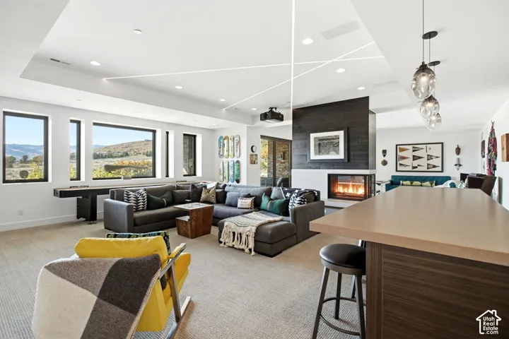 Open-concept living space featuring a recessed ceiling with linear accent lighting, floor-to-ceiling fireplace with black wood-finished surround, solid-surface kitchen peninsula, and large windows with landscape scenery