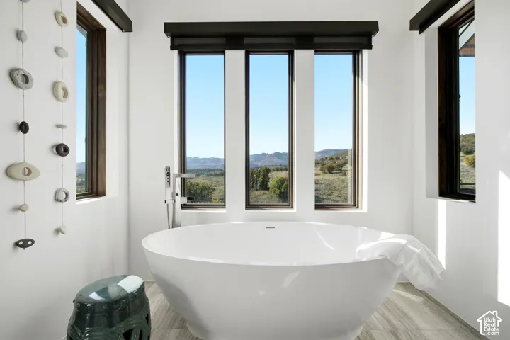 Freestanding soaking tub with a floor-mounted faucet, complemented by tall vertical windows with dark frames, wood-finish flooring, and mountain scenery