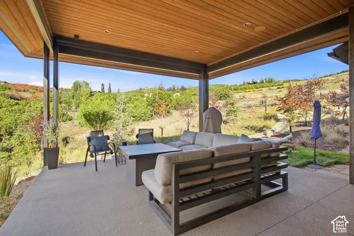 Covered concrete patio featuring a wood-paneled ceiling, recessed lighting, and metal structural supports, set against a backdrop of hillside terrain