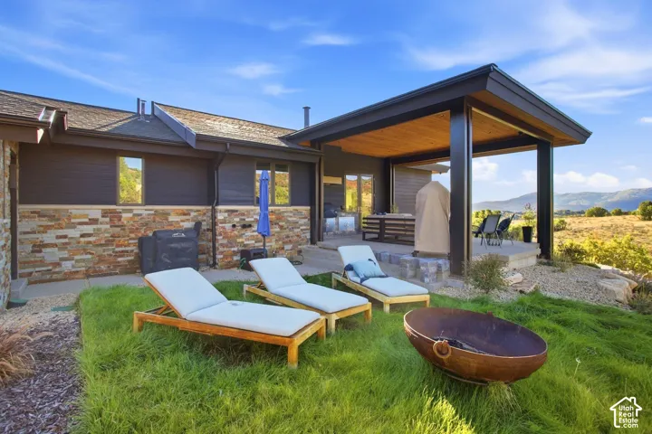 Exterior featuring stacked-stone and dark horizontal siding, a covered patio with exposed wood ceiling beams, a concrete patio foundation, and surrounding mountain scenery