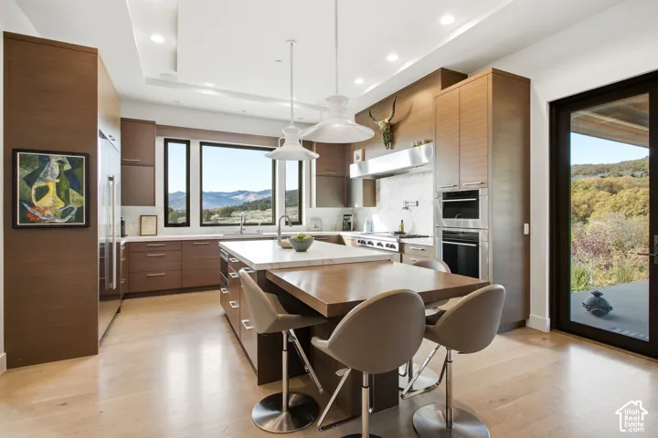 Kitchen featuring custom wood-finish cabinetry, a waterfall-edge center island with a built-in counter, stone-surfaced countertops, built-in stainless steel double ovens, and large-format windows