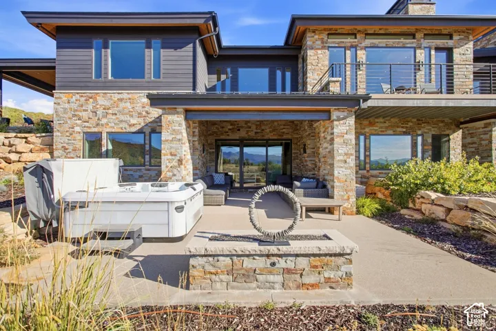 Contemporary residence featuring natural stone veneer exterior, expansive rear concrete patio, integrated stone-clad fire feature, and elevated metal-railing balcony