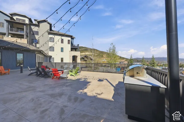 Expansive concrete terrace featuring an outdoor kitchen with a built-in stainless steel grill, mountain scenery, and overhead bistro-style lighting