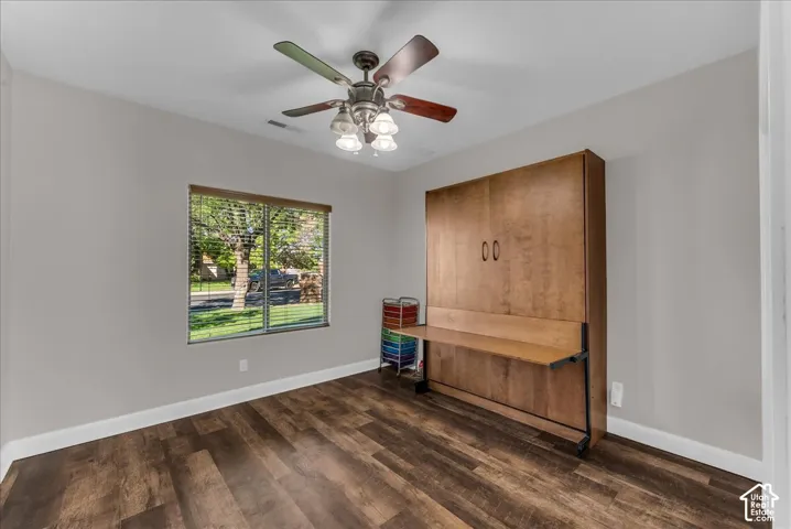 Room featuring wood-finish flooring, built-in wooden cabinetry with integrated desk surface, ceiling fan with four-light fixture, and large window with blinds