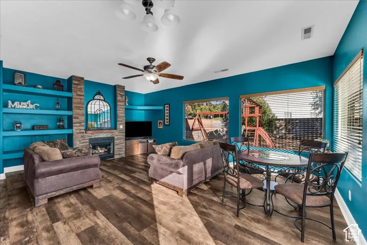 Living area featuring wood-finish flooring, a stone-accented fireplace, custom built-in shelving, a vaulted ceiling with a central fan, and large windows with horizontal blinds