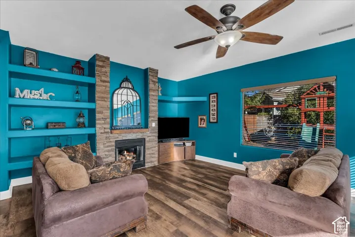 Living space featuring wood-finish flooring, a floor-to-ceiling stone-faced fireplace, built-in shelving, and a ceiling fan with integrated lighting