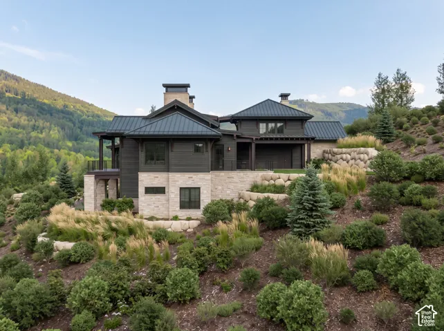 Mountain-inspired exterior featuring dark wood siding, natural stone masonry, a metal roof, and tiered boulder retaining walls set against a backdrop of mountainous terrain
