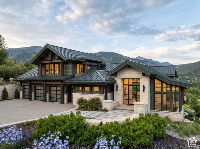 Custom residence featuring a mountain backdrop, standing seam metal roof, multi-material stone and dark wood-finish exterior, oversized glass-pane garage doors, and floor-to-ceiling windows