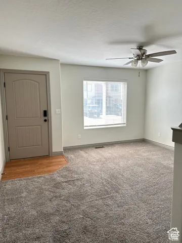 Room featuring neutral carpeting, wood-finish flooring at the entryway, a multi-panel door, window with horizontal blinds, and a ceiling fan with integrated lighting