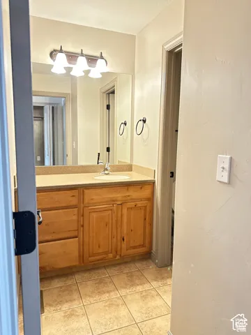 Wood-finish vanity cabinet, solid-surface countertop with integrated basin, large wall-mounted mirror, three-light fixture, and ceramic tile flooring